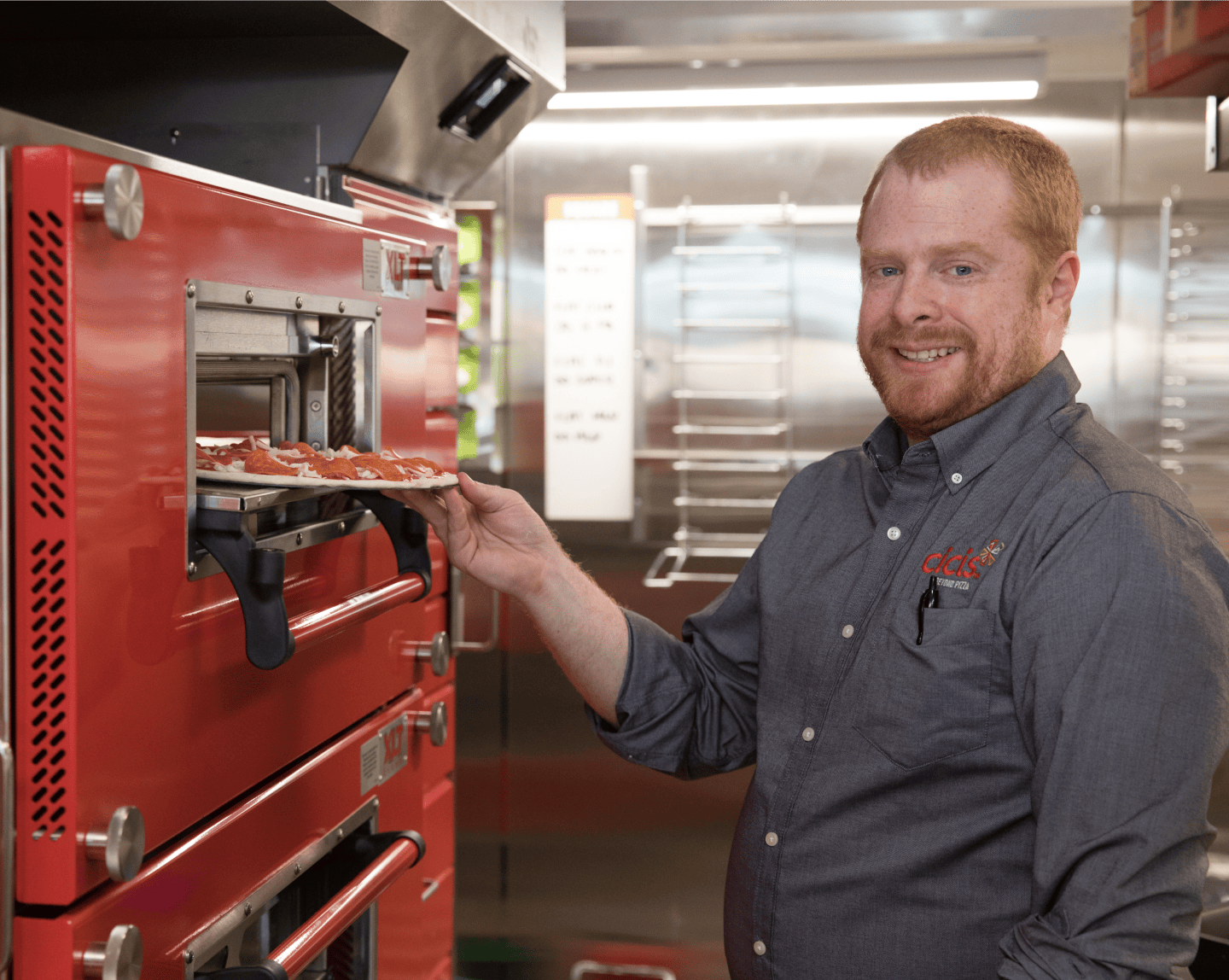 A man wearing a button-up shirt, with the Cicis pizza spark logo and text reading 'Cicis Pizza', stands in a stainless steel kitchen. He is putting a pepperoni pizza into red pizza oven.